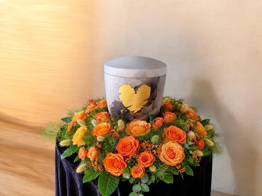 Urne mit goldenem Herz, umkränzt von orangefarbenen Rosen