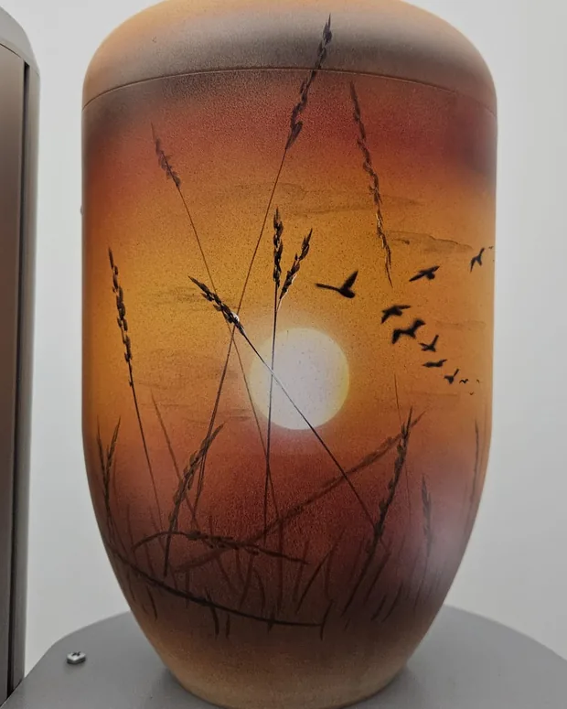 Urne mit Sonnenuntergang, davor Gräser und Vögel